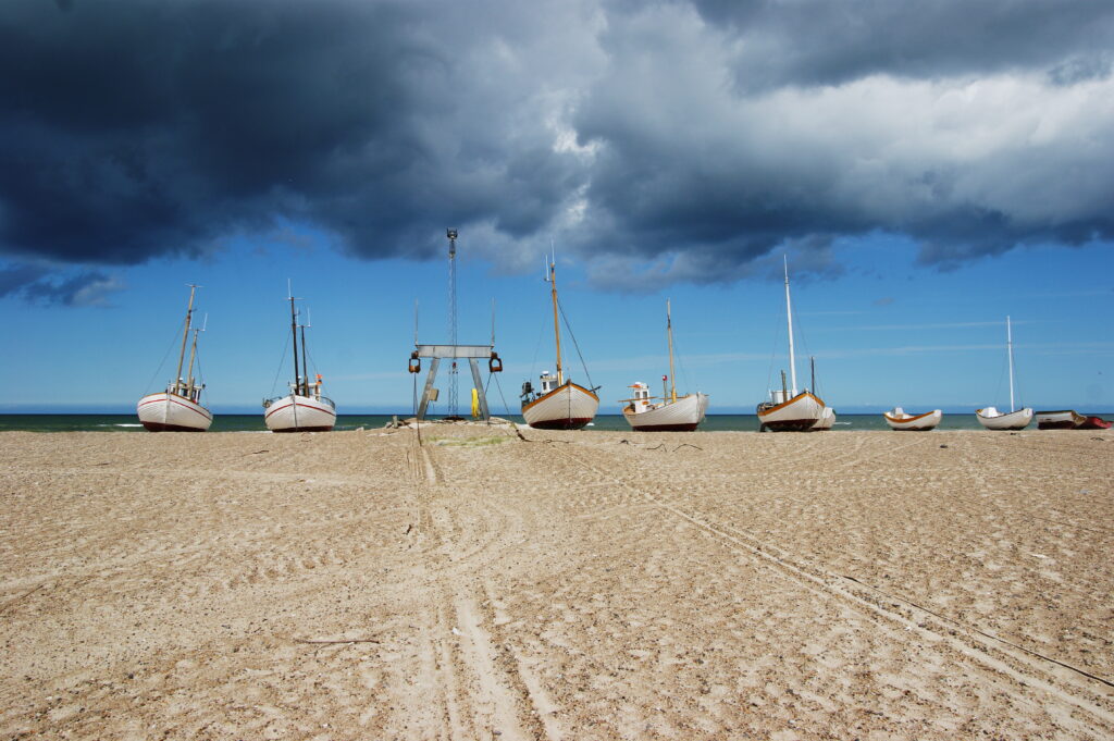 Fiskerbåde står trukket op på stranden ved Thorup Strand under dramatisk himmel med mørke skyer og blå horisont.