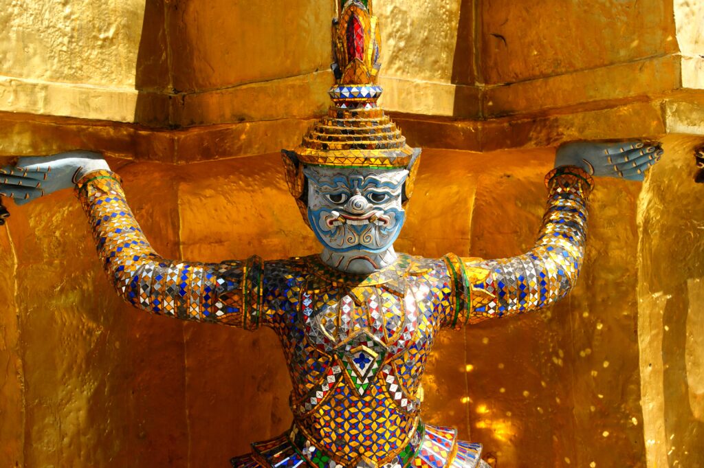 Mosaikfigur i thailandsk tempel, dækket af farvede spejle og guld, står med løftede arme og bærer templets konstruktion.