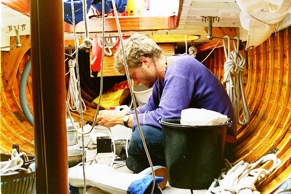 Kaptajn sidder i dørken på træsejlbåd omgivet af tovværk, træspanter og udstyr, fordybet i arbejde.