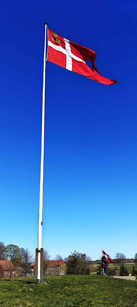 Dannebrog blafrer på høj flagstang i Jelling under en dybblå himmel.