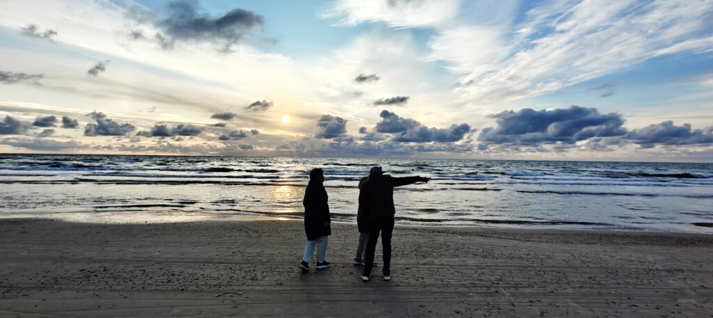 To silhuetter på en strand ved solnedgang; den ene peger ud over havet under en dramatisk, skyfyldt himmel.